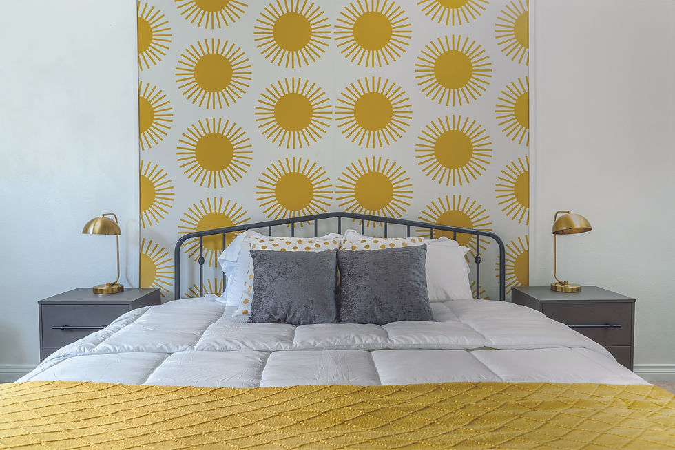 white bedroom wella, with brighton yellow sun patterned wallpaper. Metal bedframe with two white pillows, two grey square pillows. White duvet and yellow blanket at the bottom.