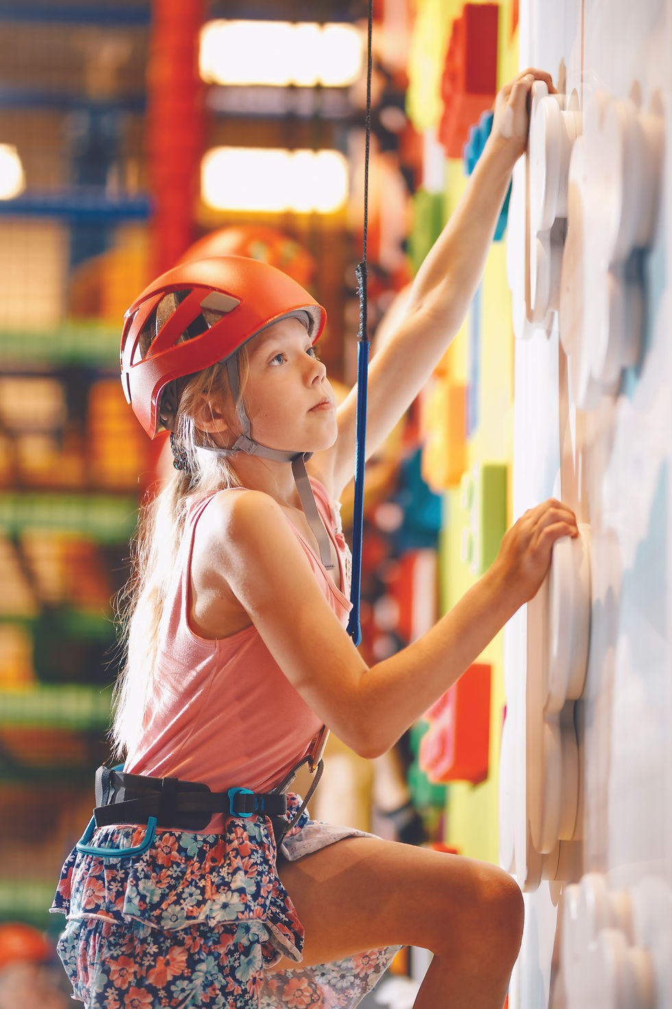 Young girl in a red helmet climbs a colorful indoor rock wall, reaching for a hold with a focused expression.