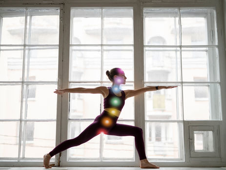 Woman in purple yoga wear holds a warrior pose in front of bright window panes, with glowing rainbow chakra lights on her body.