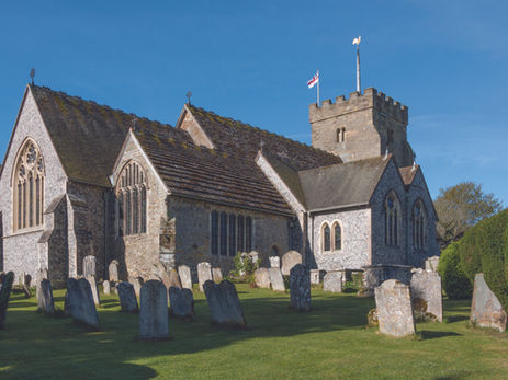 Stone church with arched windows and a tower, surrounded by gravestones on grass. A flag flies atop against a clear blue sky.