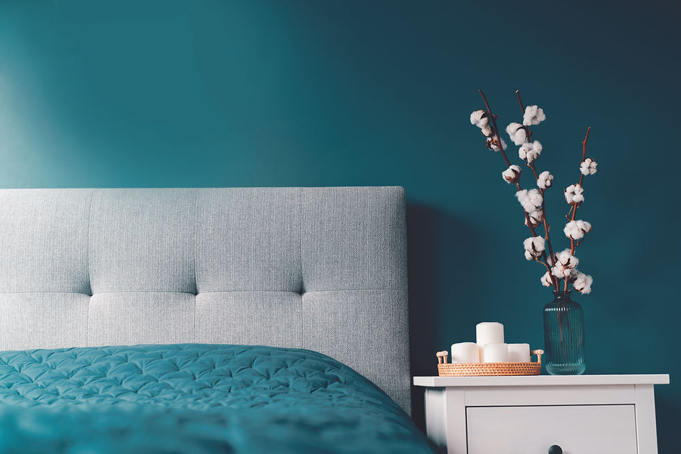 A close-up of a bedroom, with a teal coloured wall, a light grey headboard, and a teal bedspread. To the right is the top of the white side table, with 4 white candles and a glass vase with pink spring flowers in it.