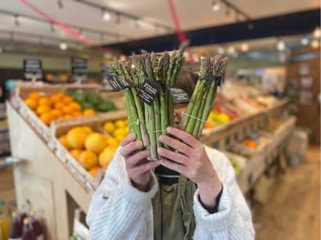Person in a market holding three bunches of asparagus labeled "Bedlam Farms." Background displays colorful fruits and vegetables.
