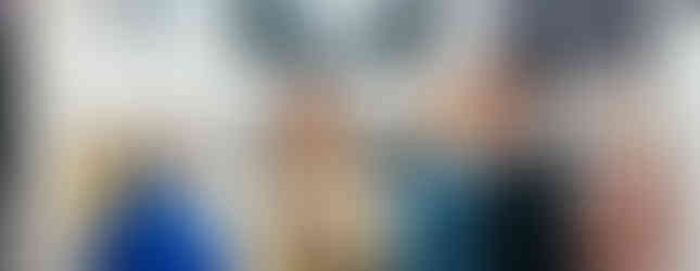 A group of people engaged in conversation at an art gallery. In the center, a woman with gray hair and a green shirt smiles while gesturing with her hands. Large-scale photographic prints of botanicals, including a dandelion and a tulip, hang on the white walls behind them.
