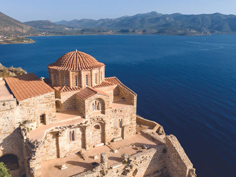 Stone monastery with red dome on a cliff above deep blue sea, with mountains and coastline under a clear sky.