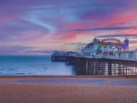 A wide panoramic shot of the Brighton Palace Pier in England at sunset. The historic Victorian pier extends into a calm blue sea under a vibrant sky filled with wispy purple, pink, and orange clouds. In the foreground, a pebbly beach stretches across the bottom of the frame, while the pier’s ornate white buildings and "Brighton Palace Pier" signage are illuminated against the twilight.