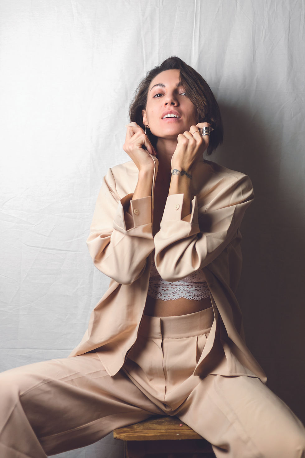 Woman in a beige suit and lace top sitting on a stool, adjusting her earrings against a plain backdrop, exuding confidence.