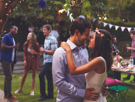 Smiling couple embrace nose-to-nose at a garden party with string lights, bunting, and guests mingling in the background.