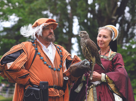 Medieval-costumed man and woman hold a falcon in a green park, both looking serious.