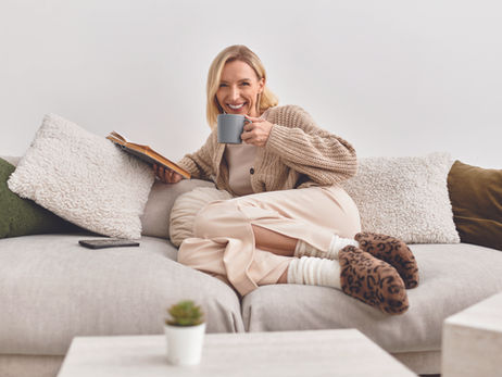 Smiling woman in a cozy sweater sits on a sofa with a mug and open book, surrounded by pillows in a bright living room.