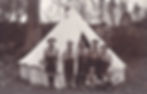 Seven young scouts in uniforms stand in front of a white canvas tent in a wooded area. Two crouch in front. Overcast sky, bicycles nearby.
