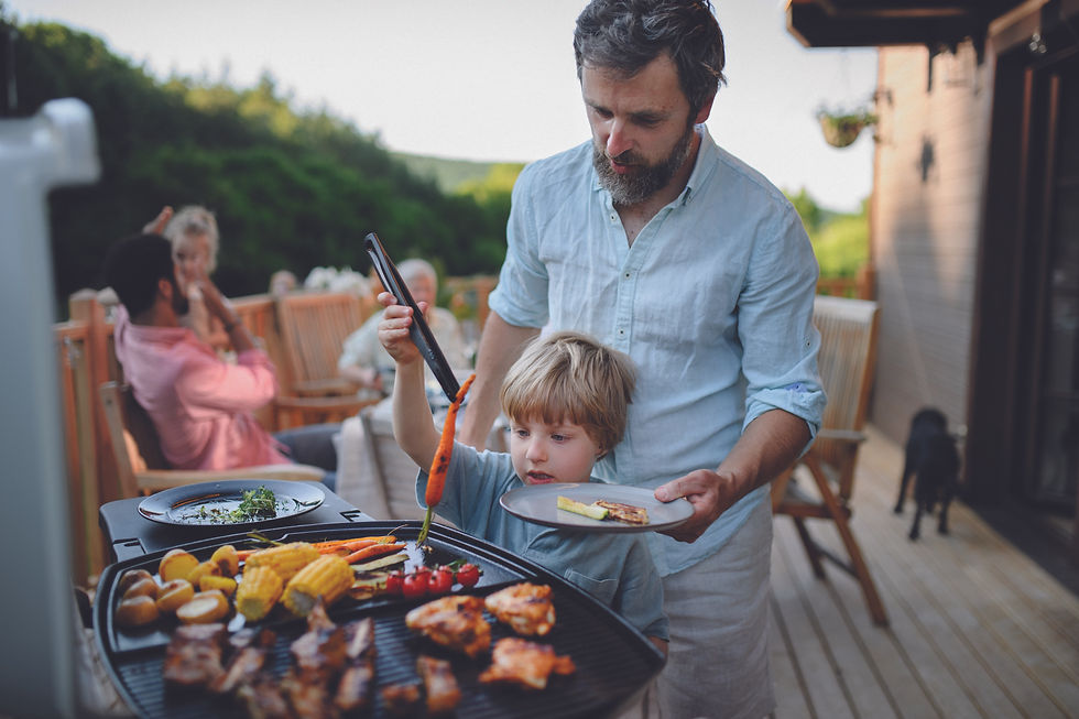 Man and child grilling food outdoors on a wooden deck, surrounded by family. Greenery in the background. Warm, relaxed atmosphere.