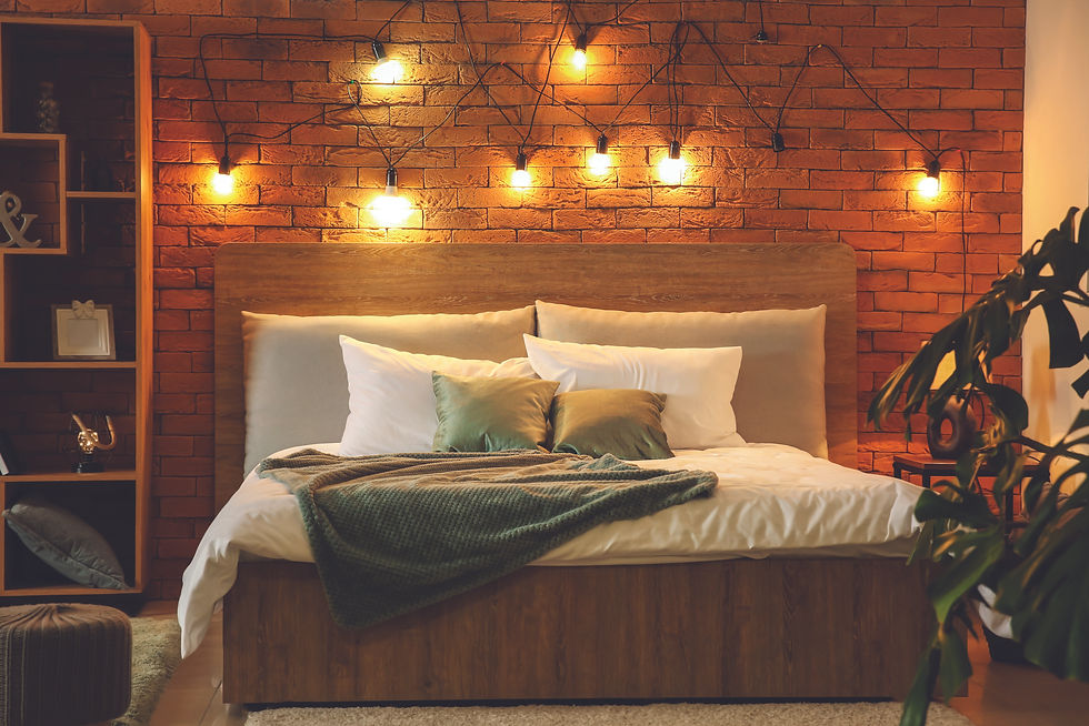 A cosy bedroom with bare brick wall, strung lights, a wooden double bec, with 6 luxuary silk pillows at the top, a white sheet, green thrown and green plant. ti the right.