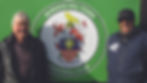 Two men smiling in front of a green sign with "Burgess Hill Town Football Club" emblem. The mood is cheerful and casual.