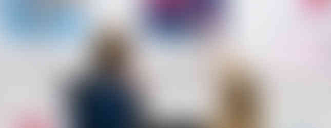 A man and a woman stand before a gallery wall featuring a series of floral paintings. The woman points toward a square canvas depicting vibrant pink poppies in a glass vase against a dark blue background. The surrounding paintings show similar floral themes in various colours.