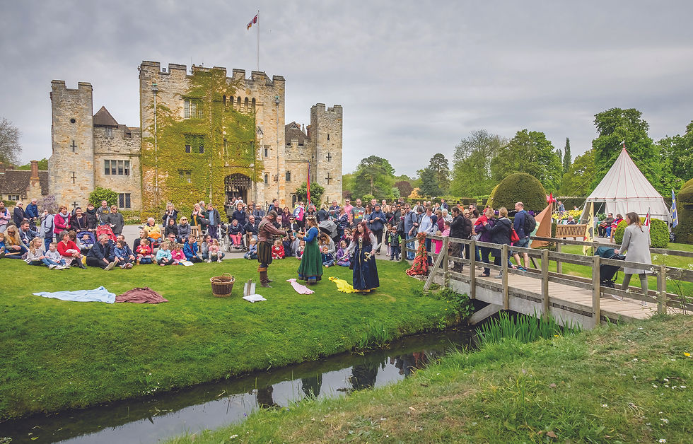 Crowd watches medieval performers on a lawn beside an ivy-covered castle, with a bridge, stream, and white tent.