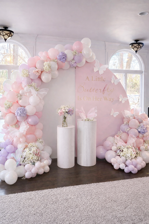 Pastel balloon arch, pink backdrop with text that says a little butterfly is on her way, pedestals, flowers