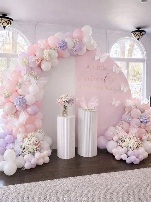 Pastel balloons in white pink and lavendar, 2 arch backdrop one white one pink that says "A Little Butterfly is On her Way", fresh flowers in the balloons, 2 white fluted pedestals one with a butterfly on top the other with a floral arrangement