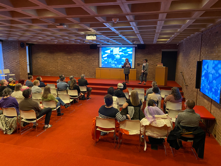 A concrete room with red carpet featuring a presentation screen hanging from the far wall. Two woman stand at the front of the image, introducing the event.
