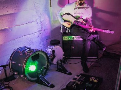 Man in white sunglasses holds a guitar labeled BILLY CAVEMAN on a stage with purple lighting