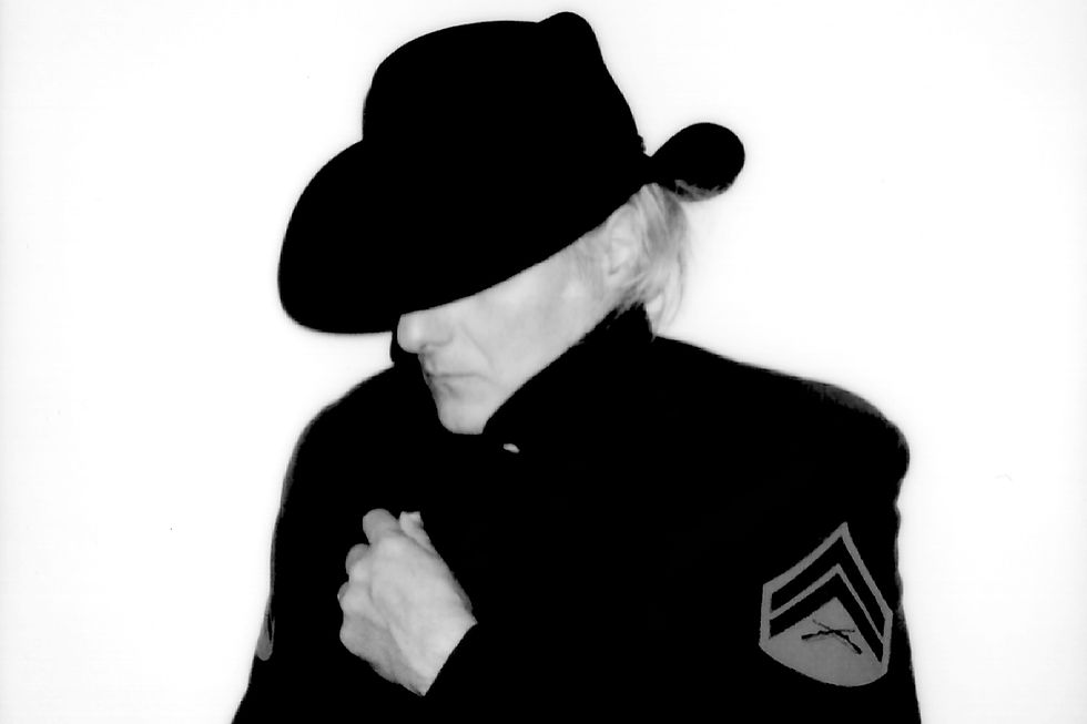 Black-and-white portrait of a person in a wide-brimmed black hat and coat, head bowed, hands clasped, with a chevron patch on sleeve.