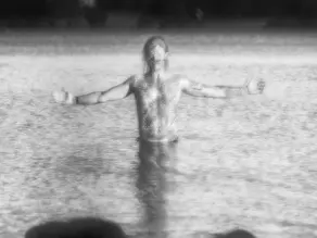 Lone person spreading his arms in the ocean through a foggy moor beside a bare tree and stone wall, creating a muted, lonely mood.