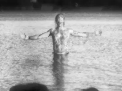 Lone person spreading his arms in the ocean through a foggy moor beside a bare tree and stone wall, creating a muted, lonely mood.