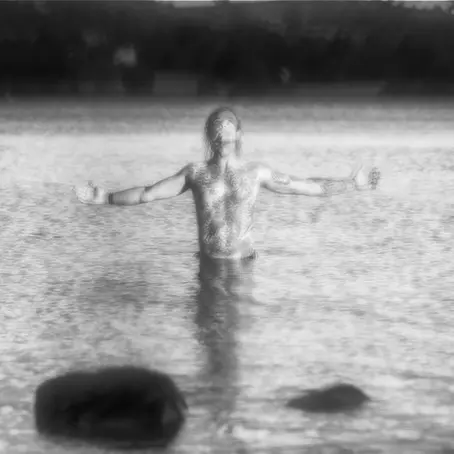 Lone person spreading his arms in the ocean through a foggy moor beside a bare tree and stone wall, creating a muted, lonely mood.