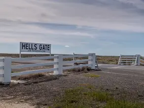 White gate and roadside sign reading HELL'S GATE on a quiet rural plain under a cloudy blue sky