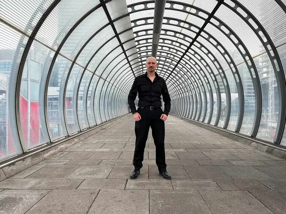 Bald man in black stands centered in a curved glass pedestrian tunnel, with city buildings faintly visible outside.