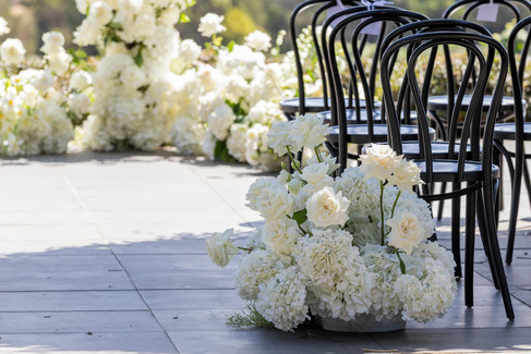 An outdoor wedding ceremony at Jackalope featuring black bentwood chairs on a stone terrace. Lush white floral arrangements frame a breathtaking view of a rolling, netted vineyard landscape.