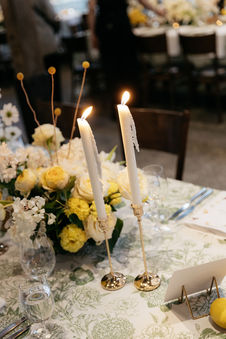 An elegant dining table is set with a white and green floral tablecloth, featuring a large centerpiece of white roses, hydrangeas, and a tall lit candle in a silver candelabra. Lemons add a touch of color, and place settings with glasses and silverware complete the sophisticated look.