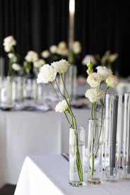 Elegant indoor reception at Jackalope featuring long tables with white linens, black-edged napkins, and bentwood chairs. Minimalist glass vases hold white roses and calla lilies beneath pleated table lamps.