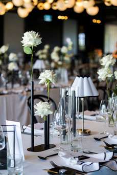 Elegant indoor reception at Jackalope featuring long tables with white linens, black-edged napkins, and bentwood chairs. Minimalist glass vases hold white roses and calla lilies beneath pleated table lamps.