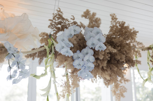 A hanging bohemian floral installation suspended from a white wood-paneled ceiling. The arrangement features feathery dried brown grasses, clusters of light blue orchids, and white calla lilies cascading downwards.