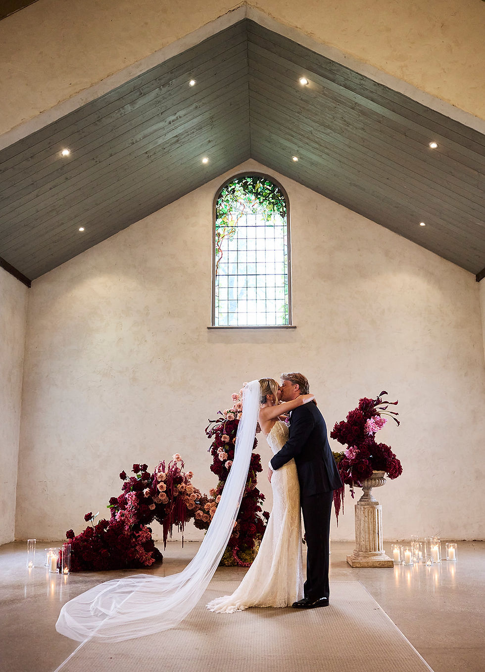 Altar view inside a rustic stone chapel looking toward the large arched window, decorated with sprawling floor arrangements of deep red, orange, and amber autumn leaves alongside flickering pillar candles.