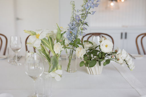 A bright, airy table setting featuring a white tablecloth, clear wine glasses, and wooden bentwood chairs in the background. The centerpiece consists of a cluster of small floral arrangements in clear glass cylinders and a white ribbed vase, displaying open white tulips, white garden roses, tall stems of light blue delphiniums, and a cascading white orchid.