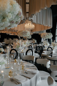 An elegant wedding reception setup featuring round tables with white tablecloths, modern white and black cushioned chairs, and tall, voluminous floral centerpieces made of white and light pink roses. The tables are meticulously set with crystal glassware, lit taper candles, and small gold perfume bottles as favors. In the background, similar tables stretch into a dimly lit room with black draped windows and a layered fabric ceiling installation.