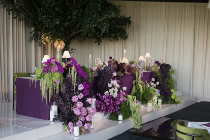 A lavish, cascading floral arrangement displayed on a stepped white platform against a solid purple backdrop. The dense, textured display features deep purple and mauve roses, vibrant purple orchids, dark purple chrysanthemums, green hydrangeas, and trailing green amaranthus. Several white pillar candles in clear glass cylinders are placed along the steps to illuminate the blooms