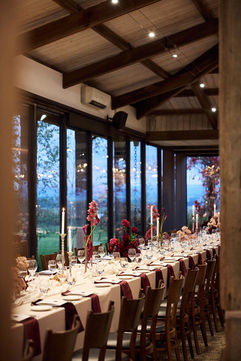 A long wedding reception table sits beside large windows at dusk. The elegant setting features white tablecloths, burgundy napkins, tall glowing taper candles, and modern pink and burgundy floral centerpieces.