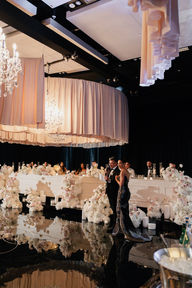 An elegant wedding reception setup featuring round tables with white tablecloths, modern white and black cushioned chairs, and tall, voluminous floral centerpieces made of white and light pink roses. The tables are meticulously set with crystal glassware, lit taper candles, and small gold perfume bottles as favors. In the background, similar tables stretch into a dimly lit room with black draped windows and a layered fabric ceiling installation.