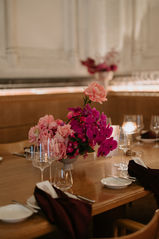 A low-profile floral centerpiece of pink roses and bright purple orchids sits on a wooden restaurant table set with wine glasses and burgundy napkins.