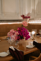 A low-profile floral centerpiece of pink roses and bright purple orchids sits on a wooden restaurant table set with wine glasses and burgundy napkins.