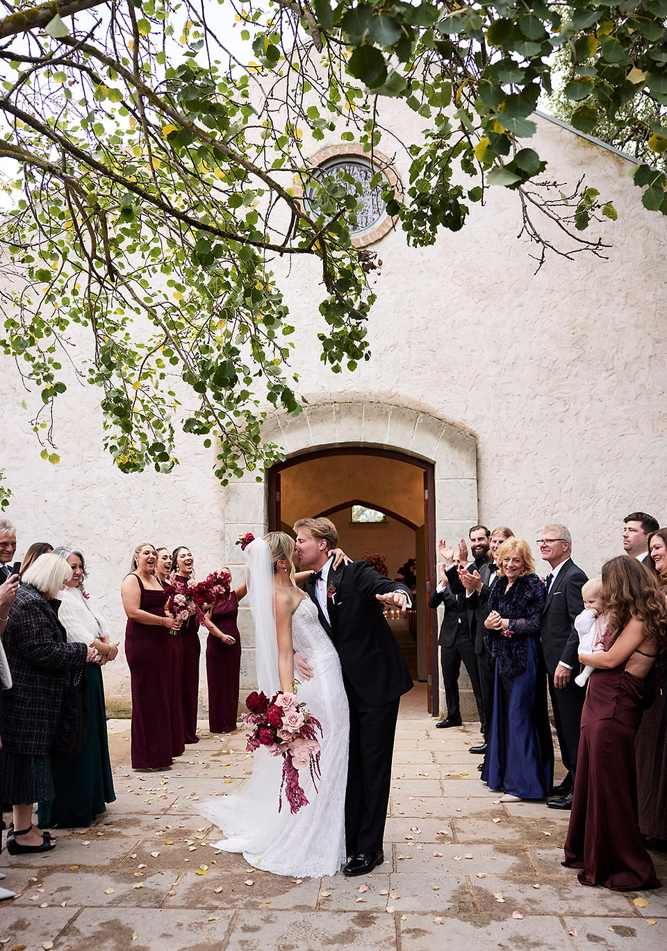A joyful newlywed couple shares a kiss while exiting a stone chapel through arched doors. Smiling guests cheer and clap on either side of them beneath leafy green tree branches.