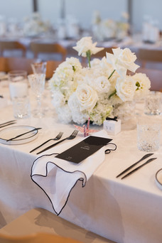 A close-up of an elegantly set wedding table. The centerpiece features a lavish arrangement of white flowers and candles. The table setting includes black-rimmed plates, black cutlery, and black menus, creating a stylish contrast against the crisp white tablecloth.