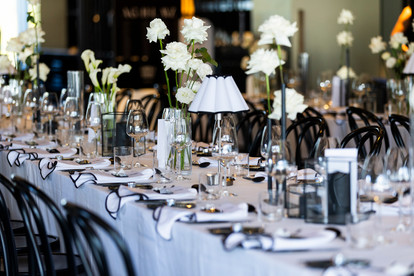 Elegant indoor reception at Jackalope featuring long tables with white linens, black-edged napkins, and bentwood chairs. Minimalist glass vases hold white roses and calla lilies beneath pleated table lamps.