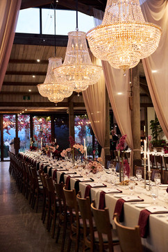 Three massive, sparkling crystal chandeliers hang over a long, beautifully decorated banquet table. Soft pink drapery, glowing candles, and striking floral arrangements complete the romantic, rustic wedding reception hall ambiance.