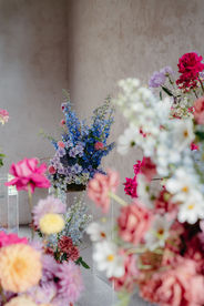 A vibrant floral arrangement featuring a variety of flowers. Prominent blooms include pink peonies, red roses, and clusters of purple and blue hydrangeas, complemented by delicate white and yellow daisies. The elevated design sits against a plain, light-colored background.