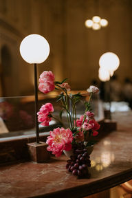 A cluster of bright pink peonies and a bunch of dark grapes sit in a small bowl on a marble counter, illuminated by soft globe lamps.