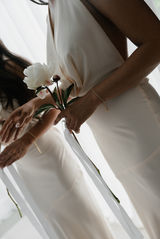 Close-up profile view of a bride and her bridesmaids from the chest down against a bright, white curtain. On the left, the bride holds a lush, full bouquet of white peonies, with a small, antique oval photo charm attached to the stems. On the right, two bridesmaids wearing elegant, cream-colored gowns hold single, long-stemmed white peonies tied with long, flowing white satin ribbons.