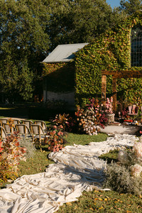 This full shot shows an outdoor wedding ceremony setup in front of a building completely covered in green ivy, with a prominent arched window at its peak. A wooden arch acts as the focal point, adorned with lush floral arrangements in shades of pink, peach, and cream. Two chairs or seating arrangements hang from the arch. More floral clusters in similar colors flank a white, flowing aisle runner that leads to the arch. Two empty chairs, matching the hanging ones, are placed on either side of the aisle in the foreground. The sky above is a clear blue.